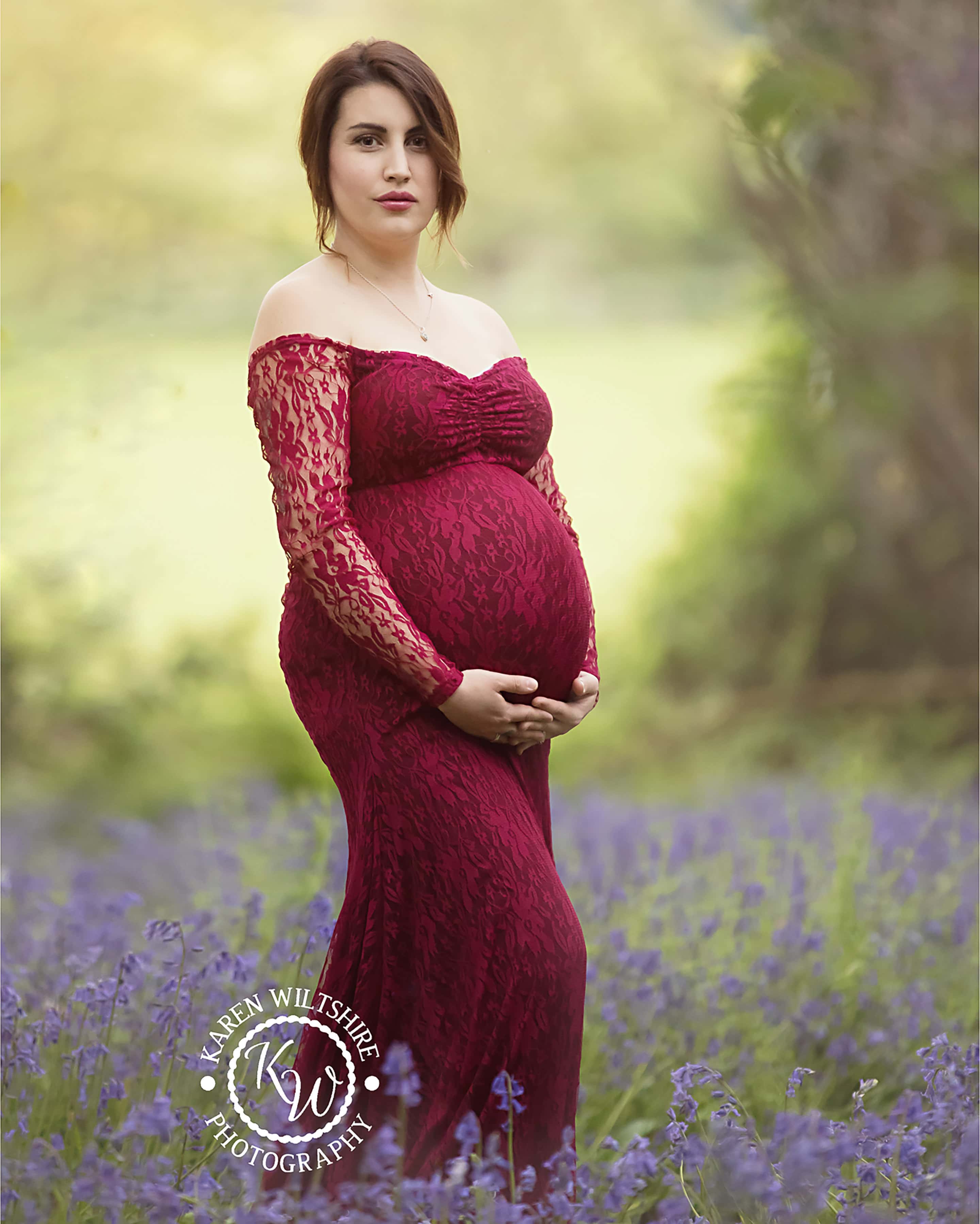 Pregnant lady in red dress standing in a bluebell wood.
